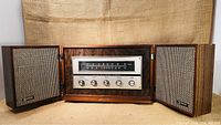 Front view of radio receiver with both speakers folded open, showing wood finish, tuning scale, five control knobs, and fabric speaker covers.
