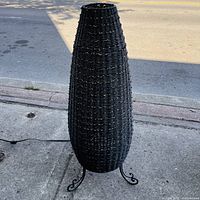 Front view of tall black wicker table lamp on sidewalk, showing woven wicker body and metal tripod base with curled feet.