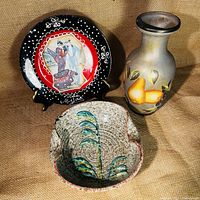 Photo of three ceramic decor items: bowl with leaf design, hand painted pear vase, and decorative plate with embroidered-style design and figurative scene.