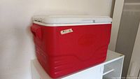 Red rectangular Coleman cooler with white lid and four cup holders, showing side view including integrated handle and wear marks.