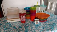 Photo of assorted cups, plates, bowls, and plastic food containers on table with blue patterned tablecloth.