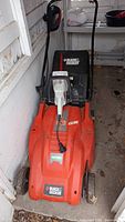 Full view of the Black & Decker electric lawn mower showing orange body, black collection bag, attached power cord, and wheels.
