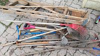 Assorted garden tools including shovels, rakes, hoes, and a wooden folding ladder placed on a paved surface, showing wear and dirt