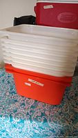 Stack of six white translucent IKEA plastic trays on top of one large red IKEA plastic storage bin on patterned blue and white tablecloth.