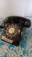 Black rotary dial telephone angled front view showing handset resting on cradle and part of coiled cord with phone resting on patterned fabric surface.