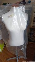 Full torso mannequin covered in clear plastic bag showing white fabric body and wooden neck cap, standing on metal base with wheels.