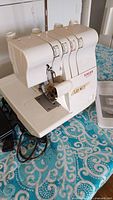 Front view of off-white Singer serger on teal patterned surface showing tension dials and presser foot