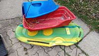 Blue, red, and green-yellow plastic sleds stacked outdoors on pavement, showing size and condition