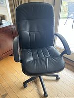 Front view of black faux leather office chair showing padded seat, backrest, armrests, and five-legged caster wheel base on wooden floor near window.
