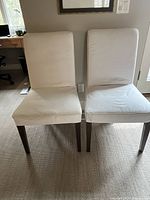 Front view of two white cotton covered IKEA dining chairs with wooden legs placed side by side on carpeted floor.