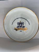 Top view of white porcelain plate showing blue and gold Queen's Plate logo with horse race imagery and text, gold rim visible.