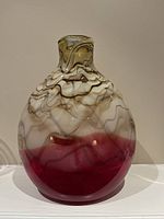 Front view of modern glass vase showing brown and beige swirling design with solid red bottom, placed on white surface against plain wall.