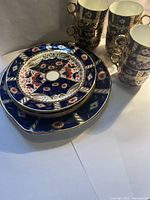 Photo showing stacked plates with intricate gold, blue, rust, and white decorative pattern and matching tea cups with gold handles stacked beside them.