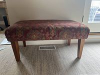 Front view of traditional wood and fabric bench with red patterned upholstery and tapered wooden legs on carpeted floor.
