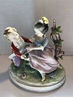 Porcelain figurine showing a couple in period dress sitting on grass with two birds on a branch beside them.