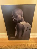 Front view of the black and white Plak-It poster displaying Vince Carter in basketball jersey, positioned against wall with wood floor beneath.