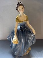 Front view of a Royal Doulton porcelain figurine of a woman in a blue dress with yellow shawl holding yellow flowers.