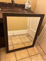 Full view of the dark brown wooden framed mirror resting against a beige wall and tiled floor, showing the entire reflective surface and rectangular frame.