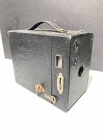 Front-left angled view of the antique box camera showing winding key, viewfinder windows, and leather handle on top
