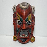 Front view of the antique wooden mask showing detailed hand carving and painting with red base color, facial features, and ornamental elements including skulls and earrings.