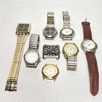Photo of seven watches and watch faces arranged on white background showing variety in band material, face shape, and color.