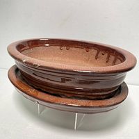 Brown oval glazed terracotta bonsai planter on top of matching water tray supported by clear rack stands.