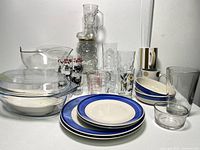 Front view of lot showing the Pyrex glass lidded dish at front, stacked ceramic plates and bowls with blue rims, clear glass pitcher, assorted drinking glasses and mugs.
