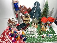 Photo showing all items including ceramic houses, artificial trees, Christmas lights, mugs, and figurines arranged on holiday-themed green and red cloths.