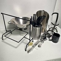 Photo showing stainless steel colander, utensil holder with black plastic kitchen utensils, metal measuring spoons still wrapped, black metal kitchen rack, paper towel holder, and black mug.