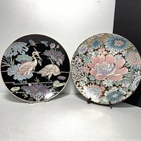 Two decorative Asian-themed wall plates side by side on stands, one with cranes and lotus flowers on black background, the other with pastel-colored floral design on gold background.