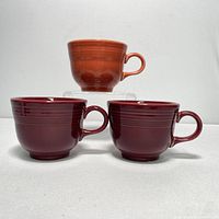 Three Fiesta coffee cups standing upright against white backdrop; two maroon cups in front, one orange cup elevated at back.