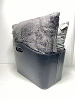 View of two modern silver-gray textured throw pillows inside a black faux leather basket with built-in handles, showing fabric texture and basket shape.