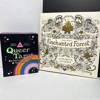 Photo showing the Queer Tarot deck in a black box with stars and rainbow design, alongside the Enchanted Forest colouring book with a detailed illustrated cover with gold accents.