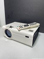 White RCA projector with lens and remote control on top, showing front and side profile.