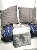 Two grey knitted look throw pillows with cable knit pattern next to a large king size pillow in original packaging and a grey plush foot warmer