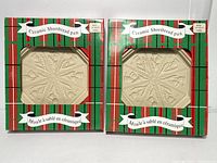 Front view of the two ceramic shortbread pans in their original green and red striped boxes with clear windows showing the beige ceramic pans inside, each divided into 8 sections with a detailed pattern.