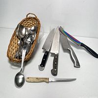 Photo showing three kitchen knives and a wicker basket with spoons and forks.