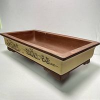 Front and side angled view showing the rectangular planter with yellow painted sides and black scenic illustrations, and brown interior and feet.