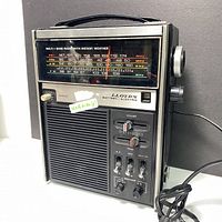 Front view of the vintage Lloyd's multi-band portable radio showing the frequency display, speaker grille, control knobs, and switches. Power cable coiled beside the device with a carrying handle on top.