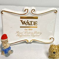 Close-up of Wade ceramic sign with gold text and decorative edges, and one small ceramic figurine of a little person wearing a red hat and blue clothing.