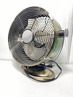 Metal table fan in metallic finish with protective grille and power cord attached, shown from a side angle.