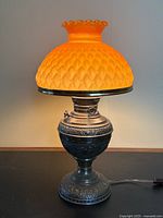 Full view of the antique brass oil lamp converted to electric, showing the glowing orange glass shade and ornate base.