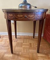 Front view of round antique mahogany side table showing floral inlay on apron, brass rim, single drawer with knob, and tapered legs on parquet floor.