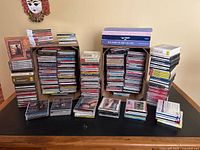 Wide view of the entire lot showing multiple stacks of CD jewel cases and boxed collections arranged on a table with a decorative wall mask above.