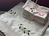 Photo showing stack of 14 napkins tied with pink ribbon alongside matched tablecloth with embroidered olive branch and decorative stitching