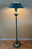 Full view of green and gold toleware floor lamp illuminated, showing entire lamp height and base detail.