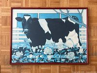 Framed poster showing stylized black and white cow with floral and fence background in blue hues, artist's name at bottom.