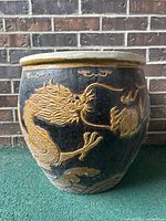 Front view showing the raised golden dragon design on a black planter against a brick wall background.