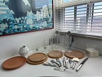 Overview of lot showing glass serving dishes on cork trivets, white ceramic vase, and various stainless steel utensils including knives and cheese servers arranged on white surface.