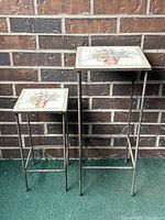 Pair of metal plant stands with square ceramic tile tops featuring hand-painted botanical design and artist's signature, placed against a brick wall.